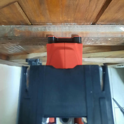 A red and black cordless tool, likely a drill or screwdriver, positioned underneath a wooden table or shelf, with a black plastic base or holder in the foreground.