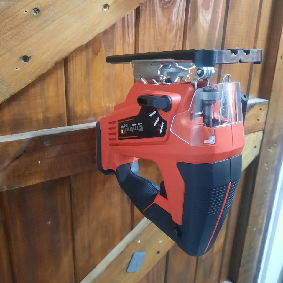 A cordless jigsaw saw with a red and black body and clear guard, hanging against a wooden wall.