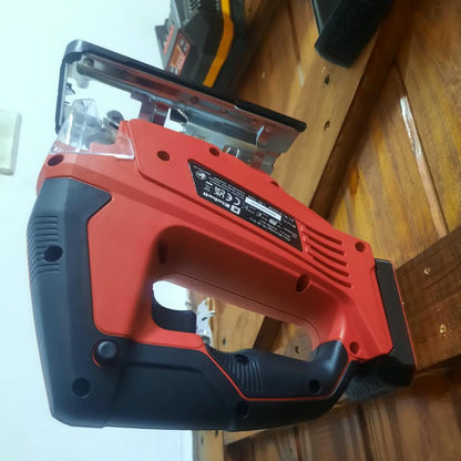A cordless jigsaw saw with a red and black body, shown resting on a wooden surface in an indoor setting.