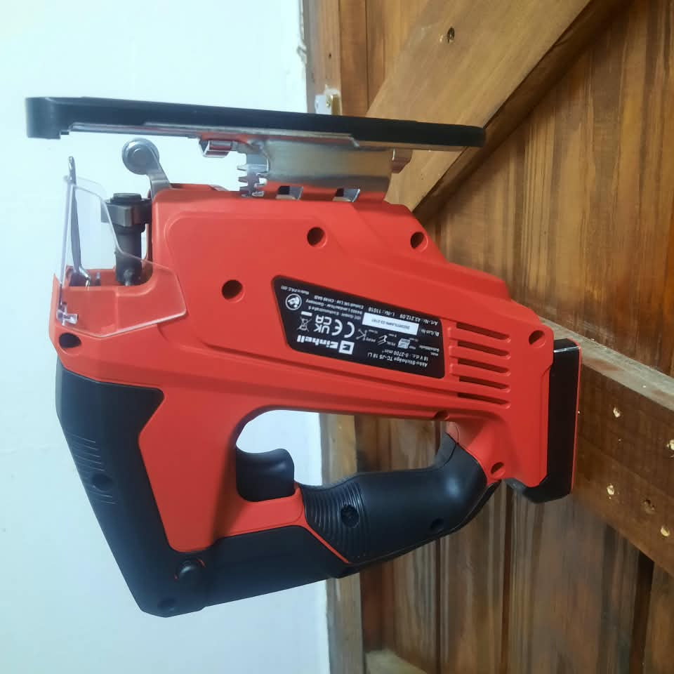 A cordless jigsaw saw with a red and black body, shown hanging against a wooden wall and white wall background.