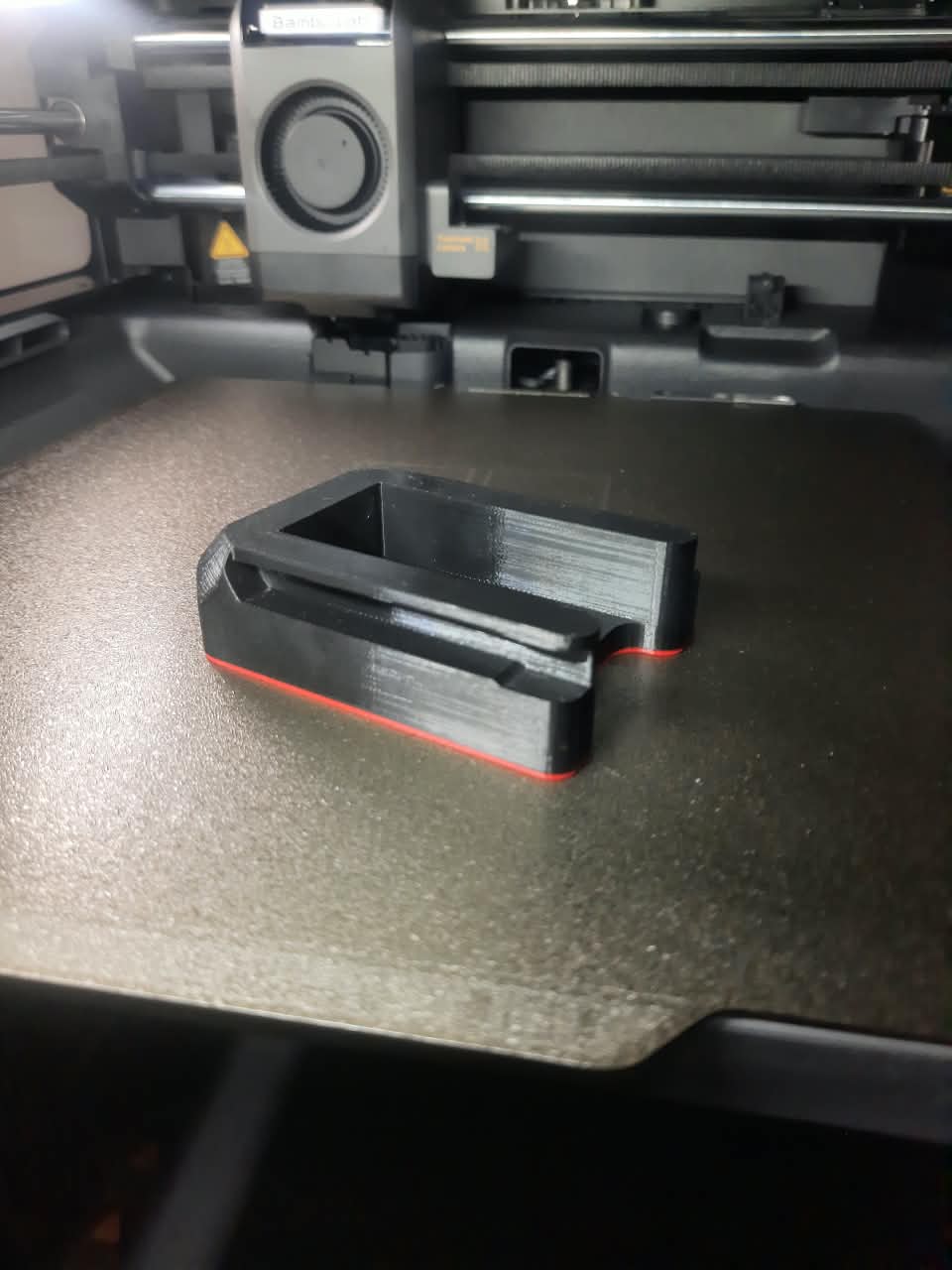 A black 3D-printed part with a thin red outline, resting on the bed of a 3D printer.