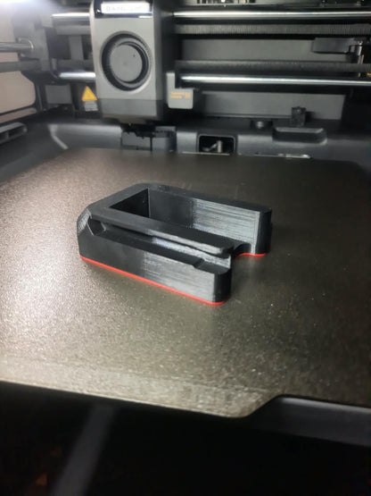 A black 3D-printed part with a thin red outline, resting on the bed of a 3D printer.