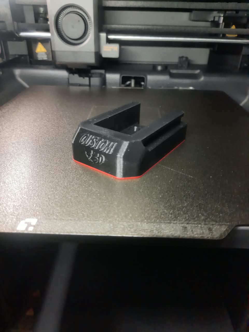 A black 3D-printed custom bracket or stand with white 'CUSTOM 3D' text and a red accent line, resting on a dark 3D printer bed.